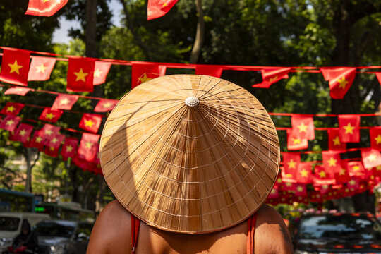 View of a conical hat obscuring the face of a person, against a backdrop of vibrant red flags with yellow stars, Hanoi, Hanoi, Vietnam.