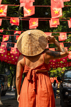 View of a woman in an orange dress and traditional hat looks up at strings of bright red flags with yellow stars, celebrating in the vibrant street, Hanoi, Hanoi, Vietnam.