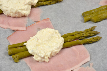 A thick slice of pink ham is topped with three glossy, preserved green asparagus spears and a large dollop of white cheese filling on gray parchment paper