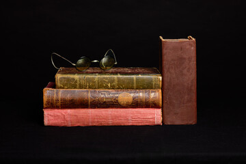 Vintage books stacked with antique spectacles and another book standing, all against a dark background, evoking a sense of history and knowledge.