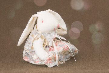 Cute handmade white plush bunny in a floral dress, sitting on a warm brown background with gentle bokeh lights.