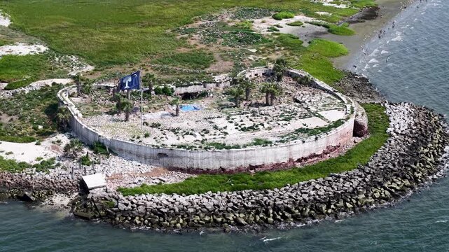 High drone view of Castle Pinckney surrounded by riprap along the coast, with visible structural ruins and lush green surroundings
