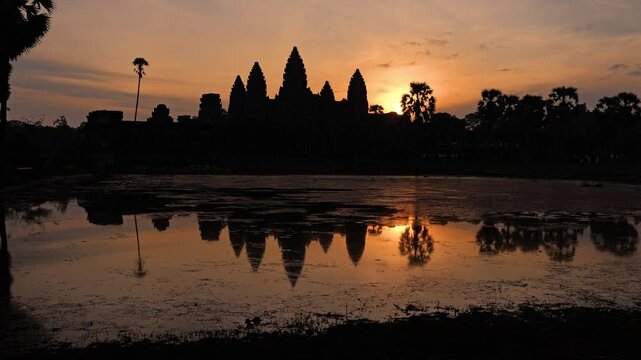 A static wide shot captures the silhouette of Angkor Wat reflected in still water as the sun rises over the temple complex in Siem Reap