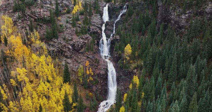 A 4K, forward tracking, close fly-over drone shot of a rushing mountain waterfall in Colorado during autumn, with ush green foliage on one side and vibrant yellow and orange fall colors on the other.