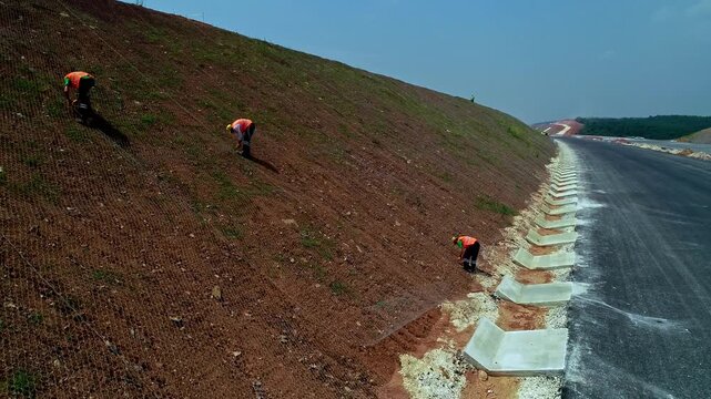 Construction workers installing erosion control and slope stabilization along a newly built asphalt road. Roadside landscaping, infrastructure safety, soil protection, and highway construction work in
