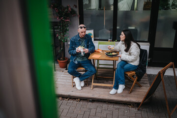 Fototapeta premium A man and a woman sit at a small outdoor cafe table, eating sandwiches. The casual scene shows friends enjoying a relaxed meal on a wooden patio.