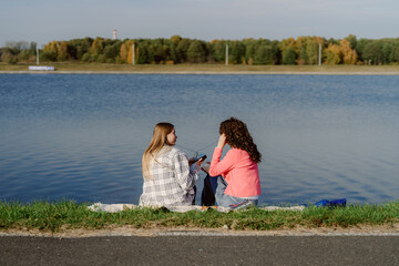 Fototapeta premium Two friends sit side by side on a blanket near a calm river, sharing stories and laughter as the sun shines on a beautiful autumn day. Their surroundings are filled with vibrant fall colors.