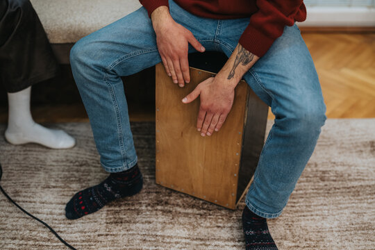 Man sits on a wooden cajon drum, hands resting on the instrument in a living room. A relaxed, casual scene with jeans, a red sweater, and visible tattoos.
