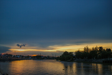 Golden sunset over Mae Klong river in Ratchaburi, Thailand. Dramatic orange sky reflected in calm water with tree silhouettes. Peaceful provincial evening landscape for travel and nature themes.