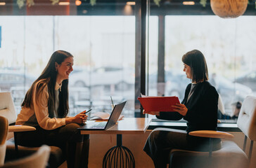 Two colleagues chat in a bright cafe, one smiling as she works on a laptop while the other holds a red binder. The scene conveys collaboration, casual business talk, and creative exchange.