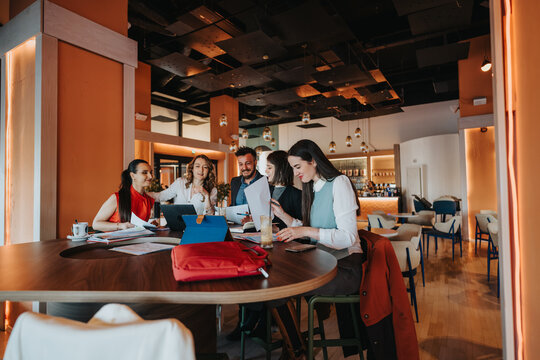 Colleagues gather around a round table in a bright cafe, collaborating on laptops and documents. Coworkers share ideas and plans in a casual, creative setting.