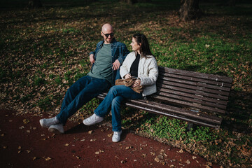 A man and woman sit on a wooden park bench, chatting casually. Autumn leaves cover the ground as they enjoy a relaxed moment outdoors.