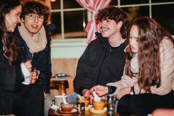 A group of friends sits together, smiling around a warm, candlelit table. The casual gathering feels friendly and festive indoors during a winter evening.