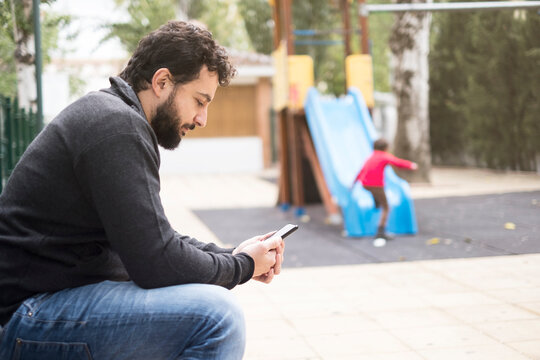Distracted father with the phone while son plays on the slide in playground