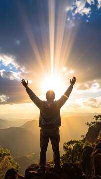 Person stands arms raised atop a rocky peak, sun bursting through clouds in golden light