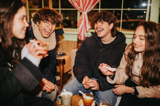 A group of young friends enjoys a lively moment, smiling and talking around a table. Warm cafe setting with mugs and snacks, capturing friendship and casual hangout.