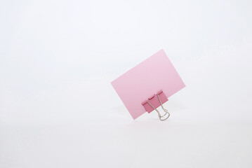 A pink note card secured with a metal binder clip on a white background. The scene is minimalistic and focuses on office supplies.