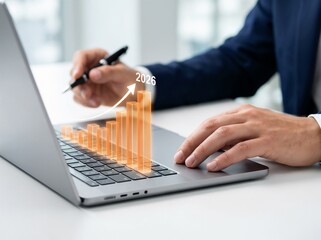 Professional hand holding pen near laptop, glowing orange 3D bar chart rising with arrow pointing to year 2026, financial growth forecast, future business strategy, success milestones, profit analysis