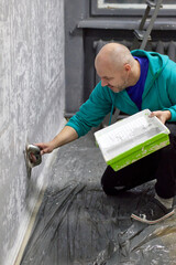 Focused bald man in a turquoise hoodie applies decorative plaster on a gray wall with a trowel, expertly crafting a textured surface indoors for a home improvement project, paint tray.