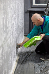 Close-up of a focused man applying textured plaster to a wall with a green tray and trowel, illustrating home improvement, interior design, and plastering skills in a renovation project.