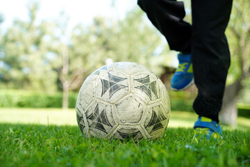   Close-up of a child's feet in blue sneakers playing with a worn soccer ball on green grass.