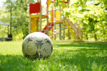 Old worn soccer ball sitting on vibrant green grass with a blurry colorful children's playground in the background.