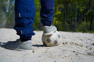 Close-up of a person's feet in grey sneakers resting one foot on a soccer ball in sand.