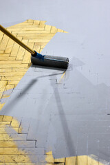 Applying gray paint to a herringbone parquet floor with a paint roller, demonstrating a modern and stylish renovation project suitable for interior design and home improvement enthusiasts.