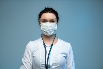 Female doctor in mask and protective glasses with stethoscope standing confidently on blue background