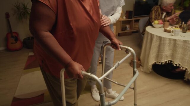 Slowmo shot of middle-aged Caucasian female caregiver supporting senior African American woman using walker while other residents relaxing by chessboard and tea in cozy nursing home