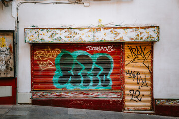 Urban street art showcasing colorful graffiti tags on a red and yellow metal roller shutter of a closed store in Calle Capitulares. Cordoba. Andalucia. Reflecting themes of abandonment and city life