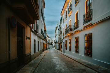 Fototapeta premium Empty narrow street with white buildings, wrought iron balconies, and glowing windows, capturing the serene evening ambiance of a historic Andalusian city in Cordoba, Spain