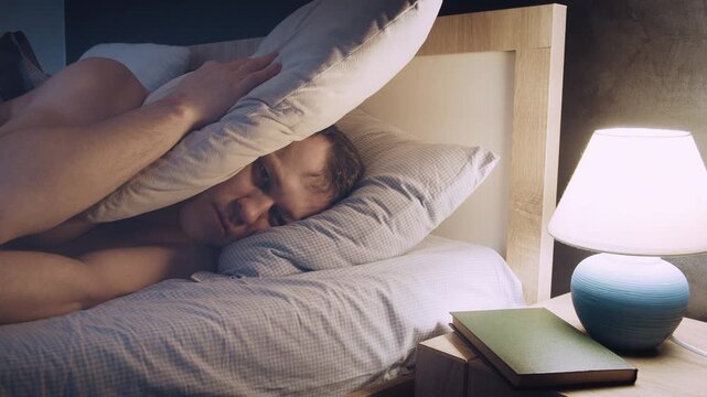 Man in Bed Covering Ears with Pillow under Lamp Light
