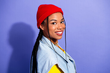 Young woman wearing a red beanie denim vest and yellow top smiles at the camera against a purple background