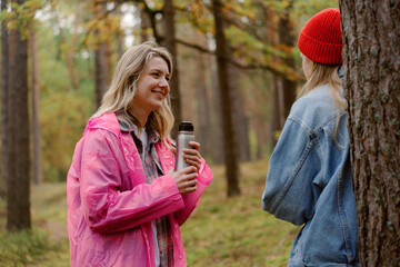 Two friends share a warm moment in a lush forest, surrounded by tall trees. The bright colors of their clothing contrast beautifully with the greenery around them, creating a cheerful atmosphere.