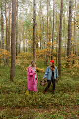 Fototapeta premium In a serene forest during autumn, two friends stroll through vibrant foliage. One wears a pink jacket and the other a denim jacket and red beanie, both enjoying a relaxing foraging adventure together.
