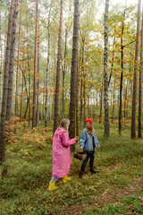 Fototapeta premium Two friends explore a vibrant forest during autumn, wearing bright outfits and gathering mushrooms. Leaves in shades of orange and yellow surround them, creating a cheerful atmosphere.