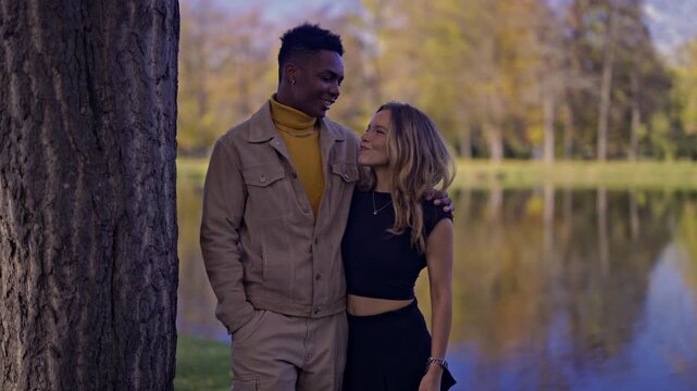 A young, diverse couple stands lovingly embraced in a vibrant autumn park. They share smiles and tender glances, showcasing their deep connection and romantic bond by a tranquil lake.