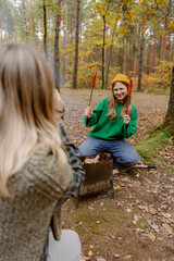 Friends gather in a serene forest during autumn, sharing laughter while cooking sausages over a campfire. Colorful leaves surround them, creating a warm, inviting atmosphere.
