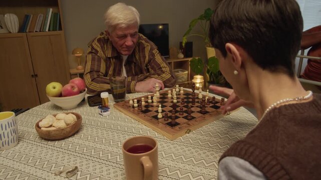 Slowmo shot of joyful seniors laughing while enjoying chess game spending relaxed evening in cozy nursing home with caregiver assisting person with mobility aid in background