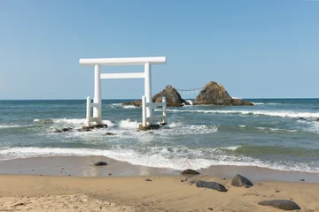 Fotobehang Zen Stenen A huge white torii gate located on the white sand beach, is lashed by the crystal-clear waves of the sea in Fukuoka, Japan. In the background, two sacred stones mark the spiritual site.  © Nina Abrevaya