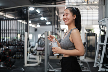 Fototapeta premium Smiling young woman in sportswear taking a water break at a modern gym. Healthy lifestyle and fitness concept.