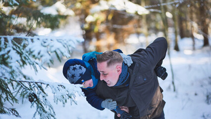 Father giving son piggyback ride, playing and having fun in beautiful snowy winter forest on sunny day