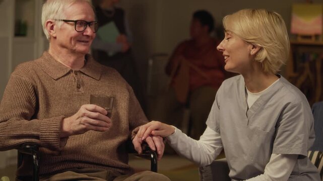 Tilt up slowmo shot of empathetic middle-aged Caucasian female caregiver listening to elderly man in wheelchair, sharing warm heartfelt conversation by evening tea in nursing home