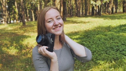 Female photographer smiling, holding camera during sunny park day, surrounded by verdant landscape and trees