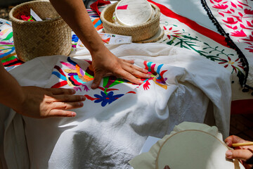 Close-up of hands designing a pattern with a pencil on embroidery fabric with a nearby traditional Mexican floral design.