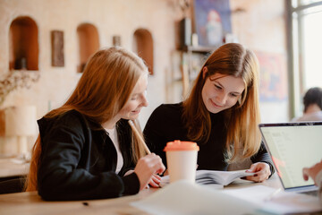 Two young women share ideas while studying in a warm, inviting cafe. Their expressions show focus and collaboration as they explore a book together, surrounded by soft lighting.