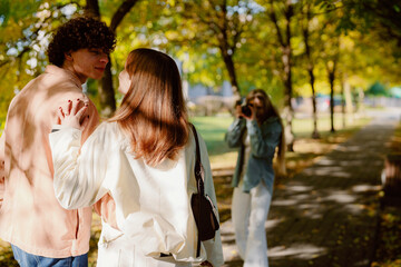 Obraz premium In a sunlit park, a couple embraces, lost in their own world, as a friend joyfully takes photos, surrounded by vibrant trees and autumn leaves creating a warm atmosphere.