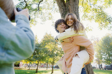 Fototapeta premium Two friends enjoy a playful moment in a vibrant park, with one giving a piggyback ride while the other captures the memory with a camera in warm sunlight.