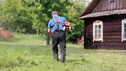 Gardener mowing grass with brushcutter near wooden house in countryside